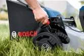 Person adjusts the height lever on a cordless lawnmower set in tall grass.