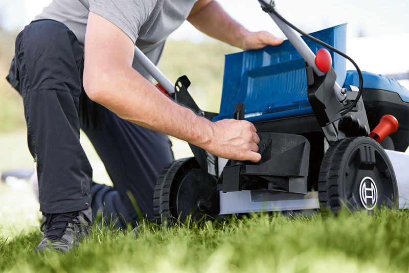A person adjusts the compartment of a cordless lawnmower on a grassy lawn.