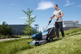 A person pushes a cordless lawnmower across a grassy slope outdoors.