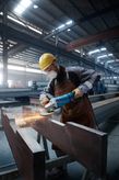 A person wearing safety equipment grinds a large metal beam, creating sparks.