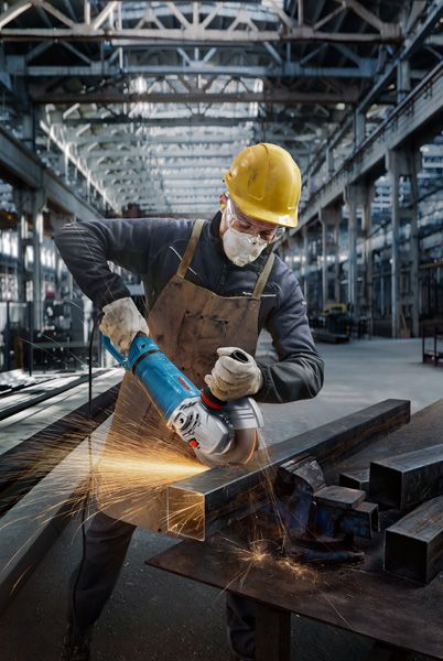 A person wearing safety equipment grinds a metal beam with an angle grinder, creating sparks.