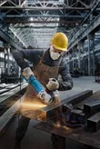 A person wearing safety equipment grinds a metal beam with an angle grinder, creating sparks.