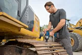 A person wearing safety equipment uses a cordless impact wrench on an excavator track.