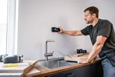 A person scans a kitchen wall above a sink with a digital detector tool.