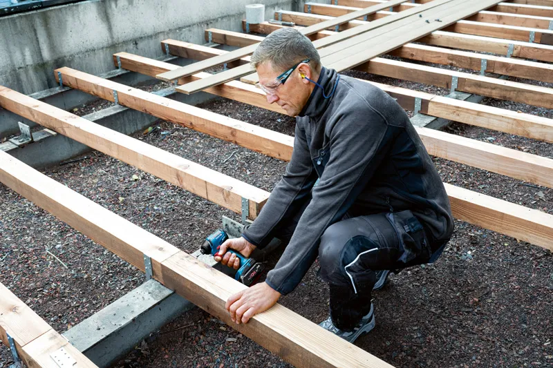 GDR 18V-220 C A person wearing safety equipment fastens wooden beams with a power drill at a construction site.