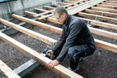 A person wearing safety equipment fastens wooden beams with a power drill at a construction site.