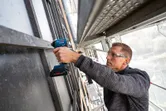A person wearing safety equipment drills into a metal frame on scaffolding.