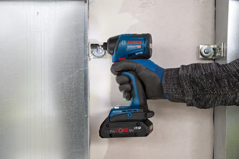 A person wearing safety equipment uses a cordless impact driver to fasten a metal bracket.