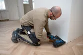 A person wearing safety equipment uses a cordless brad nailer to install baseboard trim.