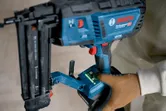 A person wearing safety equipment operates a cordless wood nailer on a light surface.