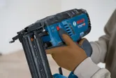 A person wearing safety equipment adjusts a cordless brad nailer.