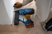 A person wearing safety equipment uses a cordless brad nailer to install trim on a floor corner.