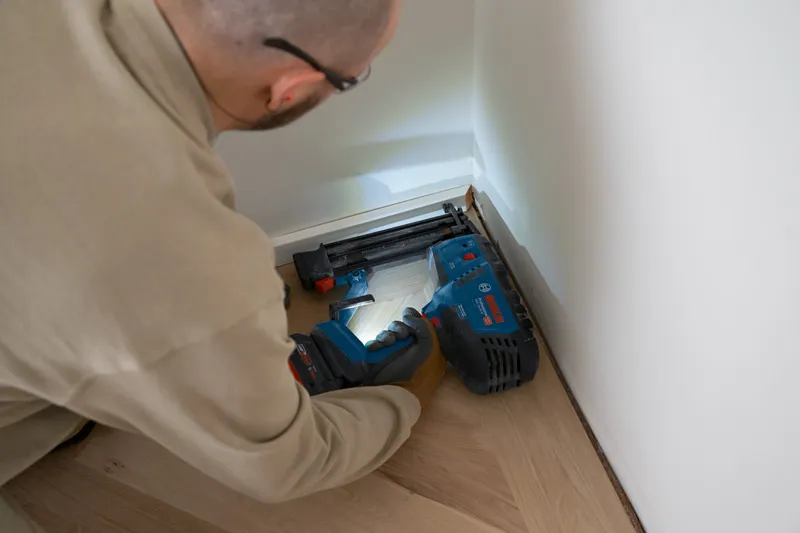 A person wearing safety equipment uses a cordless brad nailer to fasten floor trim.