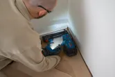 A person wearing safety equipment uses a cordless brad nailer to fasten floor trim.