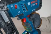 Person wearing safety equipment operates a cordless wood nailer with a gloved hand.