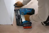 A person wearing safety equipment uses a cordless wood nailer to fasten trim to a floor.