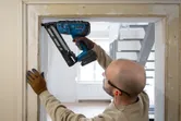 A person wearing safety equipment uses a cordless finish nailer on a door frame.