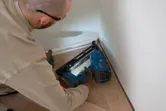 A person wearing safety equipment uses a cordless finish nailer to install floor trim.