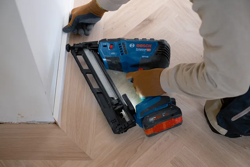 A person wearing safety equipment uses a cordless finish nailer to fix floor trim.