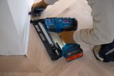 A person wearing safety equipment uses a cordless wood nailer to secure trim on a wooden floor.