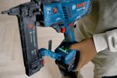 Person wearing safety equipment operates a cordless wood stapler on a wooden floor.
