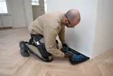 A person wearing safety equipment uses a cordless finish nailer to install floor trim.
