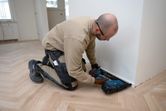 A person wearing safety equipment secures baseboard with a cordless wood nailer.