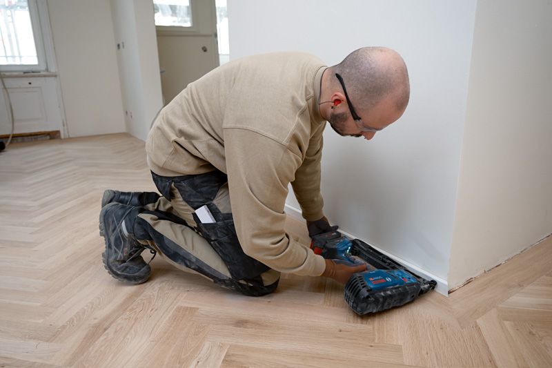 Person wearing safety equipment installs trim with a cordless wood nailer on a hardwood floor.