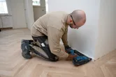 Person wearing safety equipment installs trim with a cordless wood nailer on a hardwood floor.