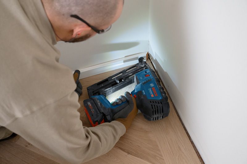 A person wearing safety equipment uses a cordless finish nailer on floor trim.