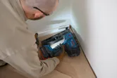 A person wearing safety equipment uses a cordless finish nailer on floor trim.