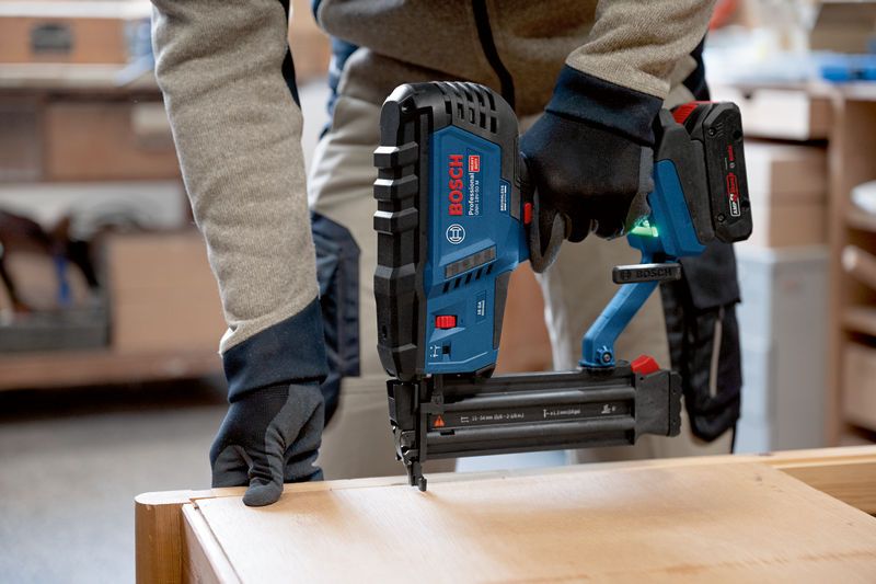 A person wearing safety equipment uses a cordless brad nailer on wooden furniture.