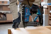 A person wearing safety equipment uses a cordless brad nailer on wooden furniture.