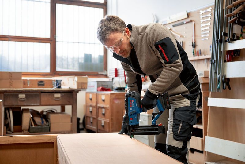 A person wearing safety equipment operates a cordless brad nailer on a wooden cabinet.