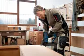 A person wearing safety equipment operates a cordless brad nailer on a wooden cabinet.