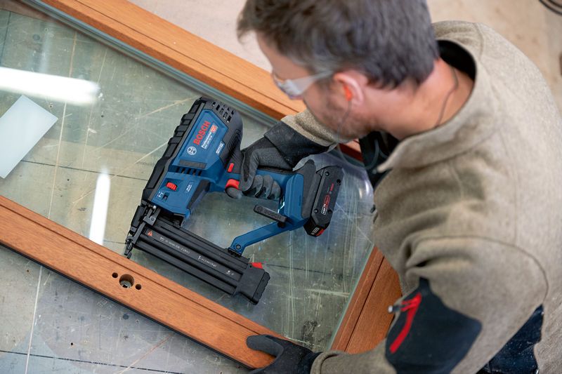 A person wearing safety equipment uses a cordless brad nailer on a wooden frame.