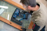 A person wearing safety equipment uses a cordless brad nailer on a wooden frame.