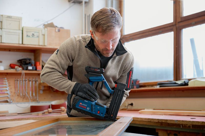 A person wearing safety equipment uses a cordless brad nailer on a wooden frame.