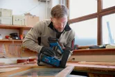 A person wearing safety equipment uses a cordless brad nailer on a wooden frame.
