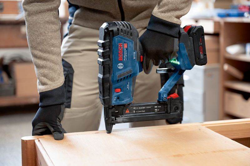 A person wearing safety equipment uses a cordless narrow crown stapler on wood.