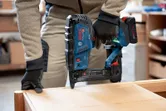 A person wearing safety equipment uses a cordless narrow crown stapler on wood.