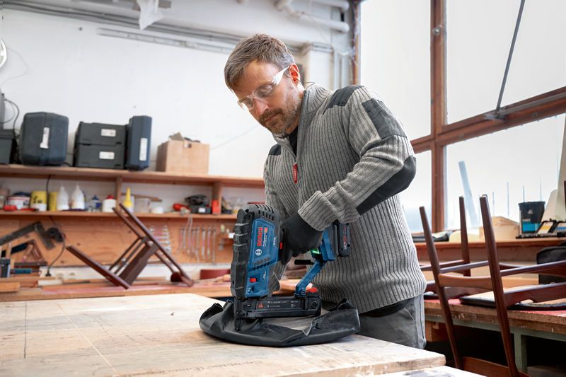 A person wearing safety equipment staples fabric to wood using a cordless wood stapler.