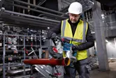 A worker wearing safety equipment uses a cordless band saw to cut a metal pipe.