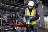 A worker wearing safety equipment uses a cordless band saw to cut a metal pipe.