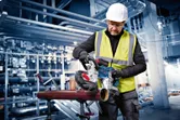 A person wearing safety equipment uses a cordless band saw to cut a metal pipe.