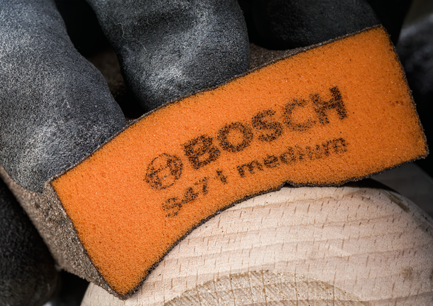Gloved hand sanding wood with an orange medium-grit sanding sponge.