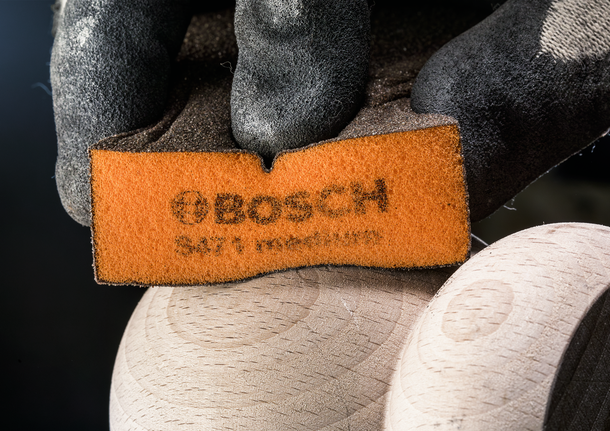 Gloved hand holding a sanding pad against a smooth wooden surface, wearing safety equipment.