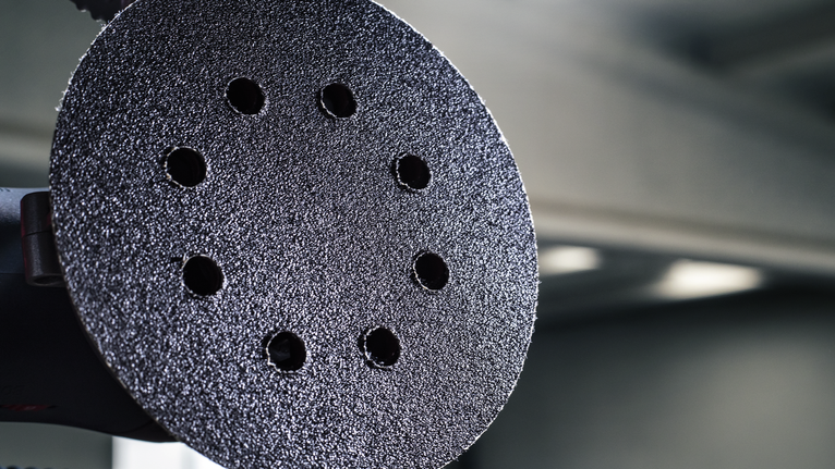 PRO F355 Sanding Disc, 150 mm, G400, 5 pcs Close-up of a circular sanding pad with multiple holes against a blurred workshop background.