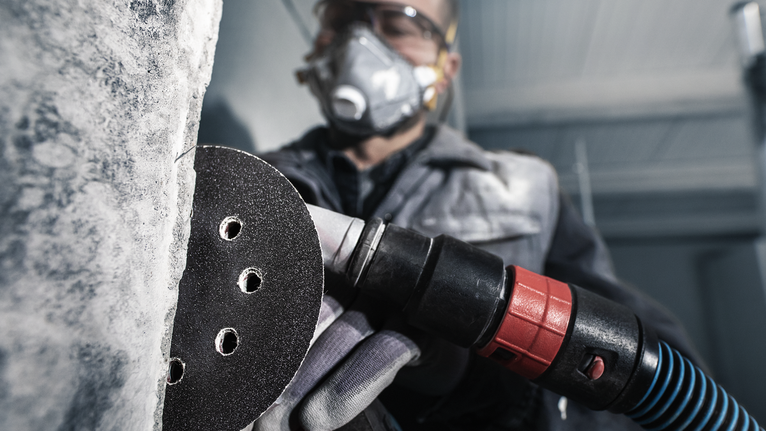 PRO F355 Sanding Disc, 150 mm, G400, 5 pcs A person wearing safety equipment sands a concrete wall with a power tool.