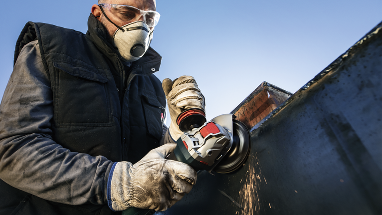 A person wearing safety equipment grinds a metal surface with a power tool.